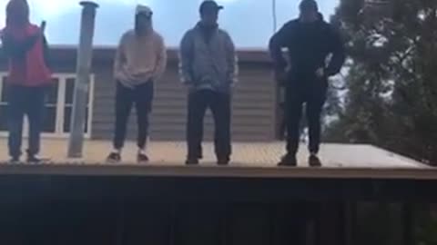 Four guys standing on roof one guy in black sweater jumps off and rolls onto grass