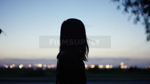 A woman turning towards lights that are off in the distance while raising her arms