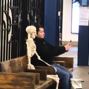 Man takes a selfie with skeleton sitting on bench in subway station