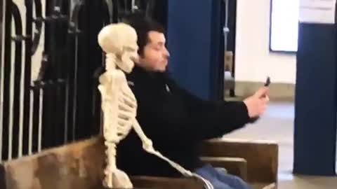 Man takes a selfie with skeleton sitting on bench in subway station