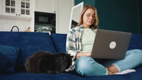 Young lady working online at home, sitting on sofa with her black cat and typing on laptop computer