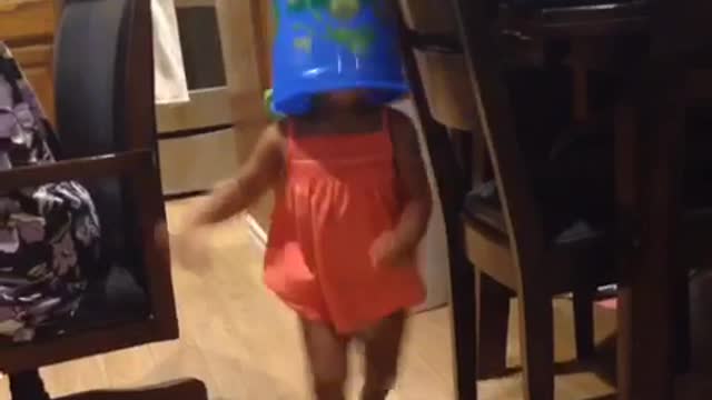 Little girl in dress with blue bucket on head walking around house runs into table