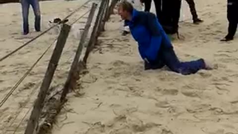 Man in blue jacket runs in beach sand tries to jump over fence