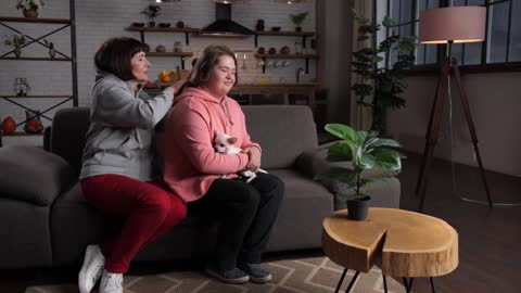 Caring female combing her handicapped daughter's hair sitting on sofa at home