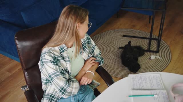 Young woman freelancer working remotely at home, taking break and looking at her black fluffy cat