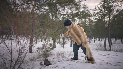 Indigenous senior hunter standing in winter forest with fox skin trying snow with stick