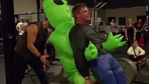 An alien does a barbell lift with a human