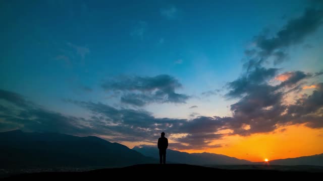 the man standing on a mountain against a sunset with northern light time lapse