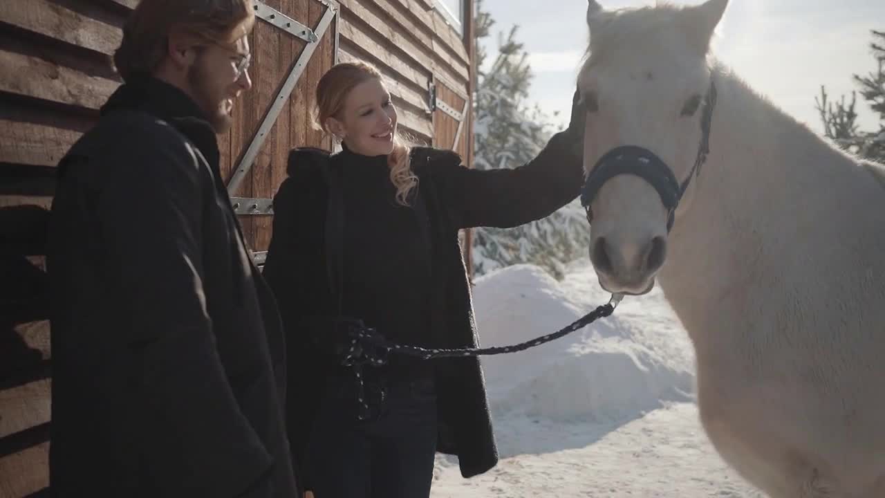 Pretty blond woman and tall bearded man standing with white horse at the snow winter ranch