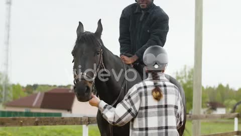 Happy Young Man Mounting, Smiling And Riding Black Horse With A Help From Young Girl