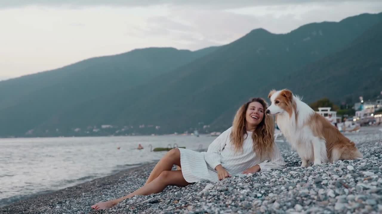 Lonely pretty woman relaxing at the beach with her dog at sunset on the background of mountain