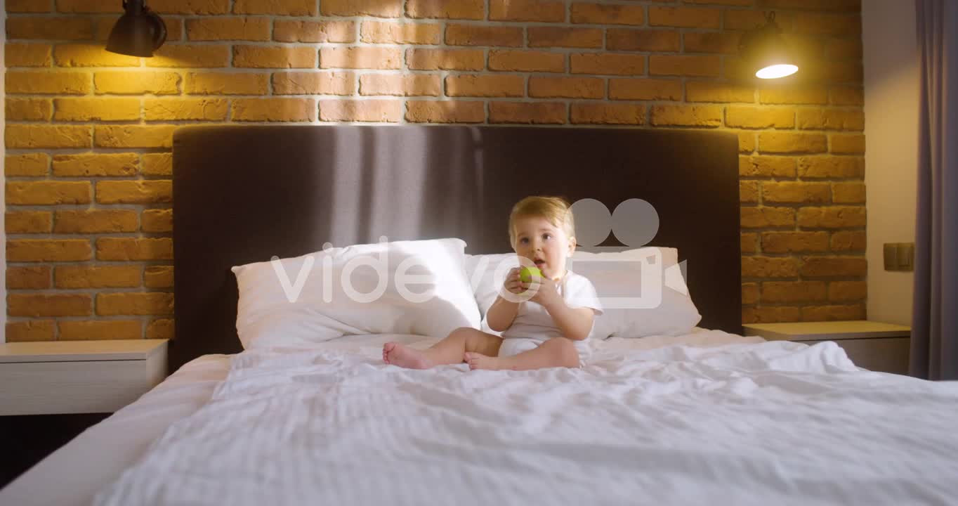 Front View Of A Baby Sitting On The Bed In Bedroom While Bitting A Green Apple