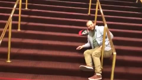 Blue shirt tries slide down gold railing on red stairs and falls