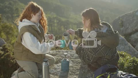 Two Young Female Hikers Drinking Hot Tea From Thermos While Sitting At Mountain Cliff On A Sunny Aut