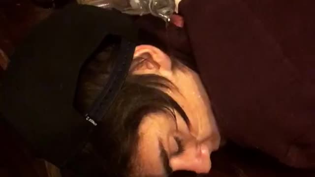 Friends pour water from coke bottle onto sleeping friends face on wooden bar