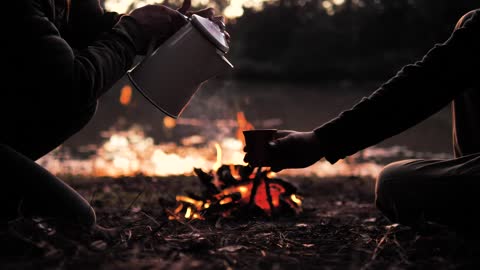 People pouring a warm drink around a campfire