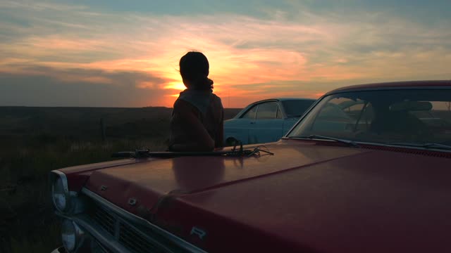 A woman leaning against a car at sunset