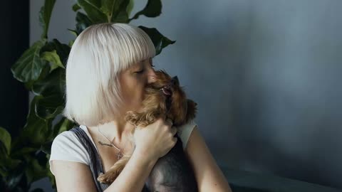 Portrait of a blonde girl kissing and hugging her little dog against a background of gray wall